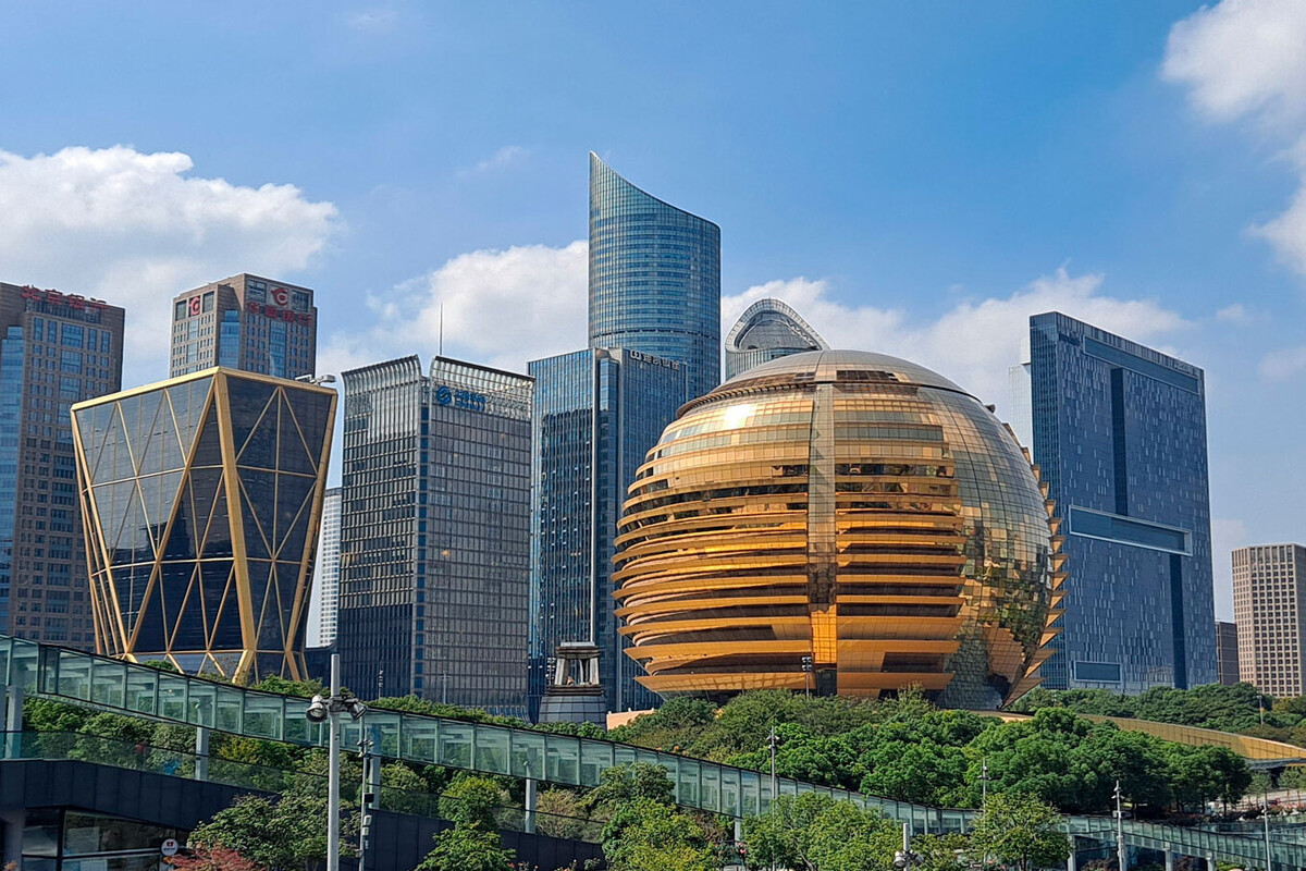 The image shows a modern city skyline featuring a variety of high-rise buildings under a bright blue sky with scattered white clouds. The focal point is a large, spherical building with a reflective golden exterior and horizontal ridges, surrounded by lush green trees at its base. Surrounding this distinctive globe-shaped structure are several tall, rectangular office buildings with glass facades, including a uniquely shaped building with a crisscross pattern of golden beams on its exterior, contributing to the city's contemporary architectural diversity.