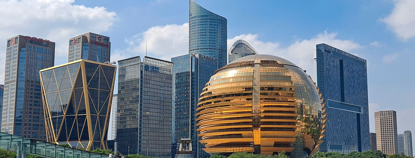 The image shows a modern urban skyline with a variety of unique and distinctive skyscrapers under a partly cloudy blue sky. In the foreground, there is a large spherical building with a reflective golden exterior, featuring horizontal band-like patterns that give it a striking appearance. Surrounding this sphere are several tall buildings with glass facades, including a pair of rectangular towers with red logos at the top, a tower with a crisscross golden framework, and other sleek high-rises with varying shapes and architectural styles, set against a backdrop of greenery at the base.