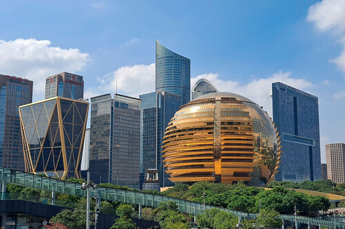 The image shows a city skyline featuring several modern high-rise buildings under a blue sky with scattered clouds. The most prominent structure is a large, spherical building with a gold reflective surface and horizontal banding, surrounded by various rectangular skyscrapers with glass facades. In the foreground, there is greenery and an elevated walkway or bridge stretching across the scene.