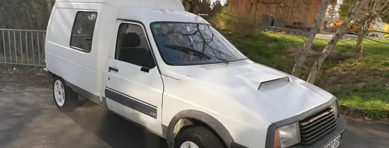 The image shows a small white van parked outdoors on a paved surface near a grassy area with trees. The van has a compact cab and an extended cargo area with a side window, suggesting it might be used for delivery or transport. The vehicle appears older, with a prominent hood scoop and black trim along the side, and it is surrounded by a natural setting with some fencing and buildings visible in the background.