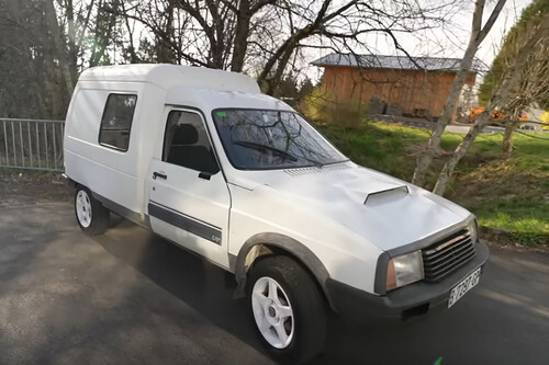 The image shows a small white utility vehicle parked on a paved surface near some trees and a wooden building in the background. The vehicle has a boxy design with a raised, enclosed cargo area at the back, featuring a small window on the side. The front of the vehicle has a simple grille and rectangular headlights, with black trim along the bottom edges and white wheels.