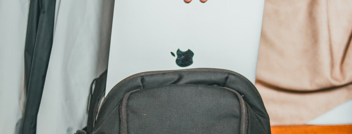 The image shows a close-up of the back of a silver MacBook laptop, partially tucked into a black backpack. The Apple logo is clearly visible on the laptop. The background includes a light-colored fabric or curtain on one side and a soft pinkish or beige cloth on the other, with a hint of a wooden surface at the bottom right corner.