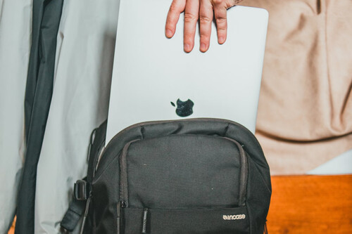 A person is placing a silver Apple laptop partially into the main compartment of a black Incase backpack. The laptop is held by the top edge with the hand visible, and the laptop's Apple logo is clearly seen. The background includes a beige surface, possibly a blanket or cushion, and the edge of a wooden surface.