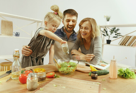 Cute Little Girl And Her Beautiful Parents Are Cutting Vegetables And Smiling While Making Salad In Kitchen At Home