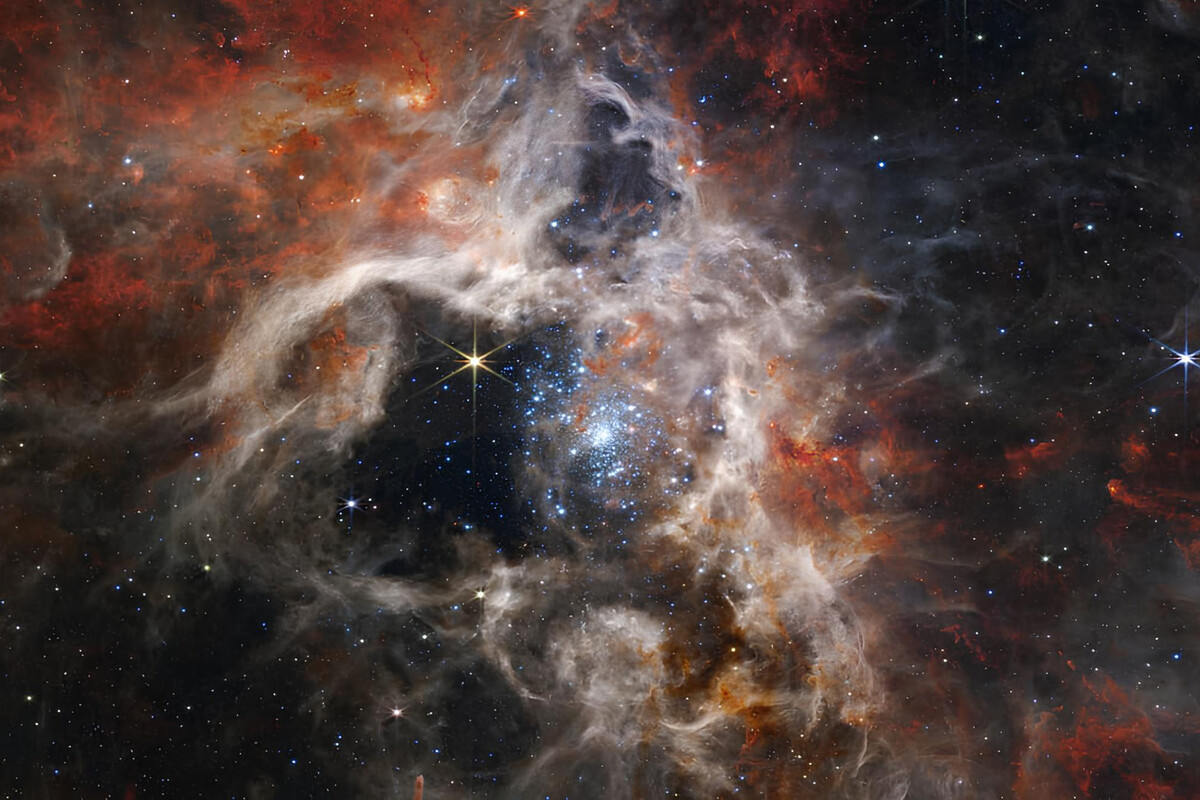 This image depicts a vibrant and dramatic cosmic scene filled with swirling clouds of gas and dust in shades of deep red, orange, and gray. At the center, a dense cluster of bright blue and white stars shines intensely, with smaller stars scattered throughout the surrounding dark space. Wispy, nebula-like textures weave around the stars, creating a sense of depth and dynamic movement against a backdrop speckled with countless twinkling stars.