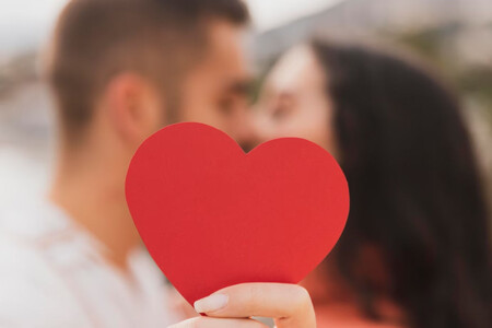 Un corazón rojo recortado aparece en primer plano, en segundo, borroso, una pareja se da un beso
