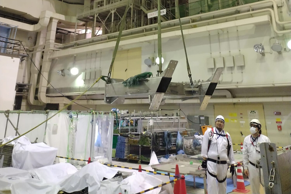 The image shows an indoor industrial or construction setting with various equipment and materials organized around the space. Two workers dressed in white protective uniforms, helmets, and harnesses stand near orange traffic cones, observing a large metal component suspended in the air by harness straps attached to an overhead crane system. The background includes metal scaffolding, pipes, cables, and machinery, indicating a controlled work environment with safety measures in place.