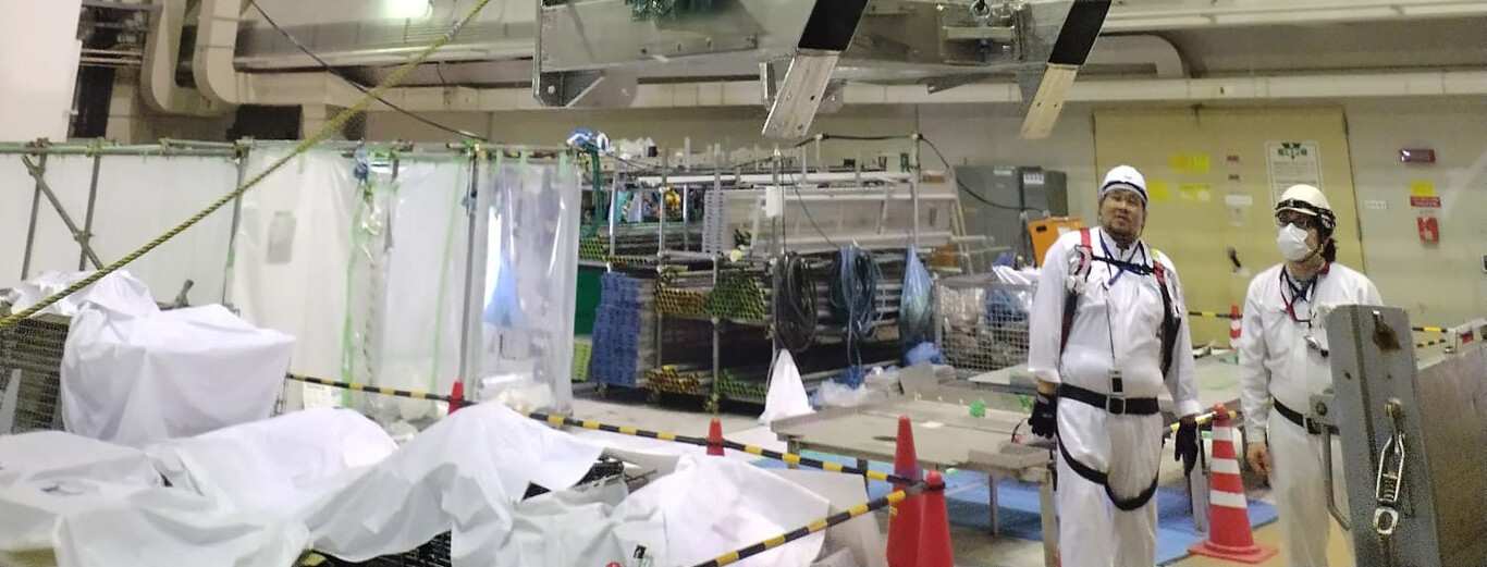 The image shows an indoor industrial or construction area with two workers dressed in white protective uniforms, helmets, and safety harnesses. They are standing near a sectioned-off workspace surrounded by caution tape and orange traffic cones. Various equipment and materials covered with white sheets are visible in the foreground, while stacks of supplies and machinery are seen in the background, all under bright overhead lighting.