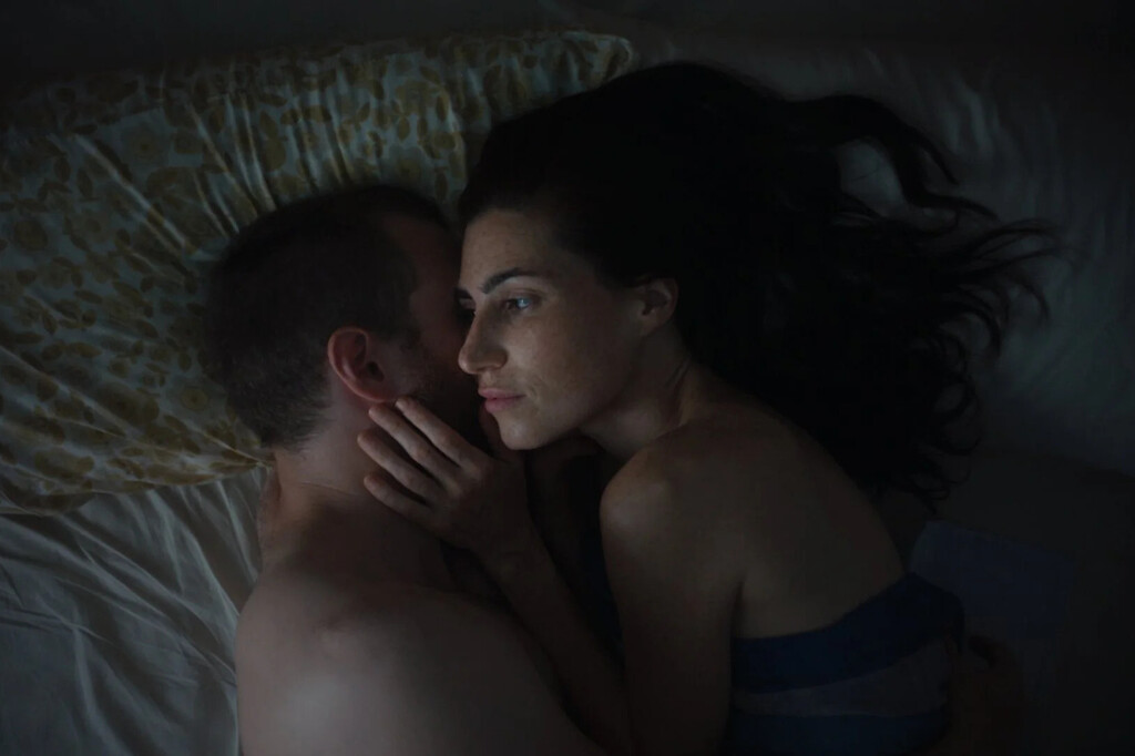 Couple in an intimate embrace, lying face to face in bed, bathed in dim light.
