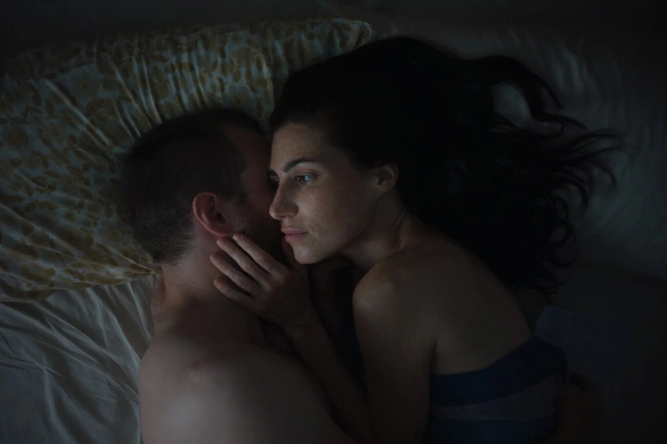 A man and woman lying closely together, the woman looking thoughtful, touching the man's neck.