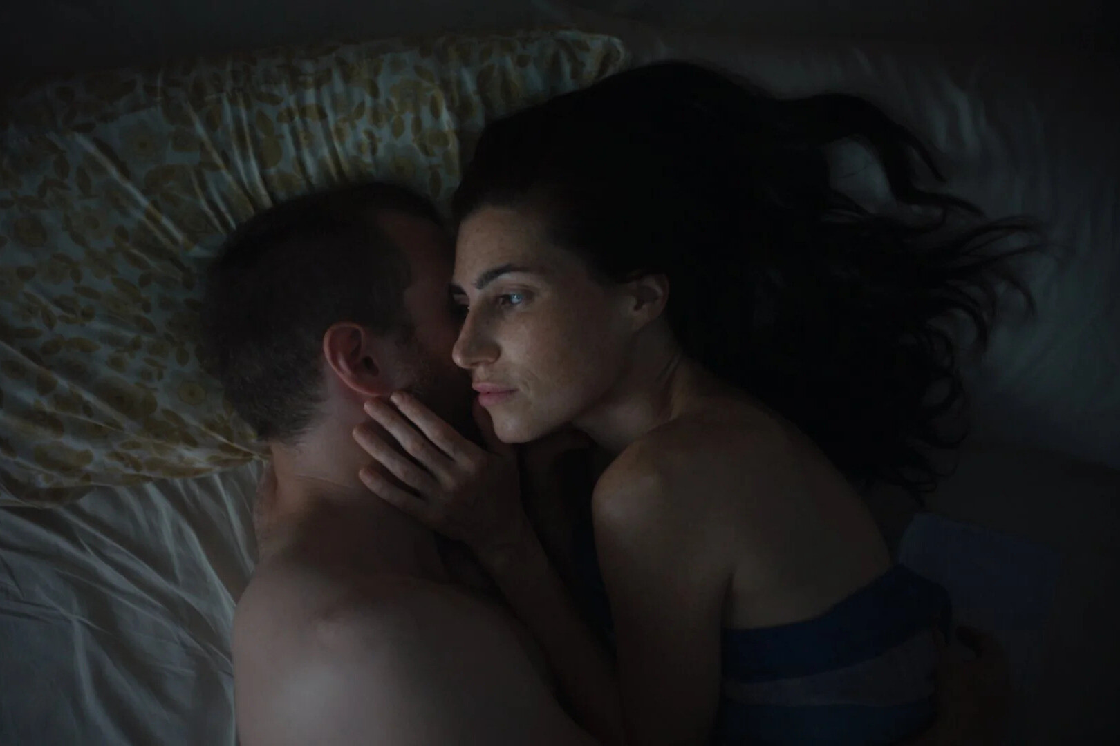 A man and a woman lying in bed close together in a dimly lit room, sharing an intimate moment.