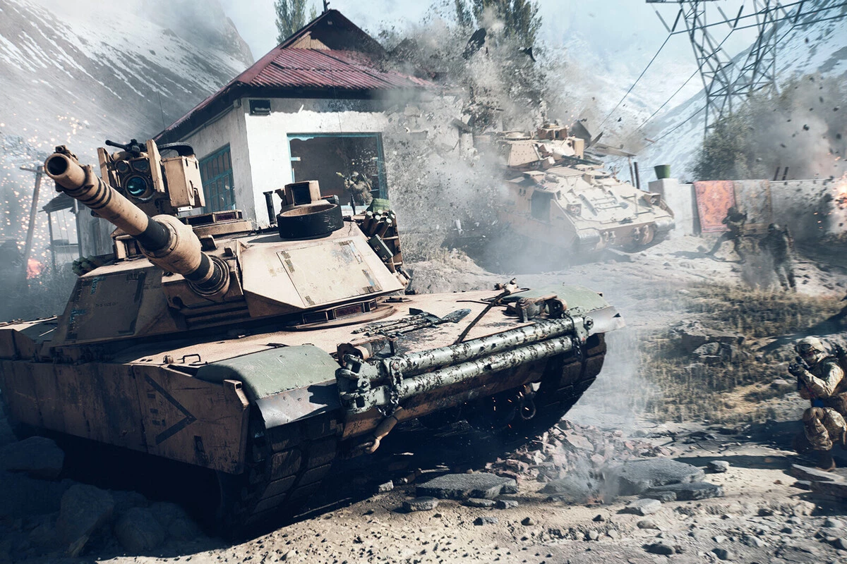 The image depicts an intense battle scene with two tan-colored tanks navigating a rocky, dusty terrain near a building with a red roof. One tank is prominently positioned in the foreground, showing wear and scratches, while the other is partially obscured by a large explosion and smoke near the building. Several soldiers in combat gear are visible, some taking cover and others advancing cautiously amid the chaos, with power lines and a mountainous background adding to the setting's rugged and war-torn atmosphere.
