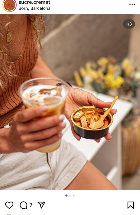 A person wearing a rust-colored ribbed tank top and white shorts is holding a glass of iced coffee with visible swirls of cream in one hand. In the other hand, they hold a small black cup filled with a caramelized dessert, likely crème brûlée, topped with a small wooden spoon and a piece of caramelized sugar. The background includes a blurred arrangement of yellow flowers and a neutral-toned wall, suggesting a cozy and inviting setting.
