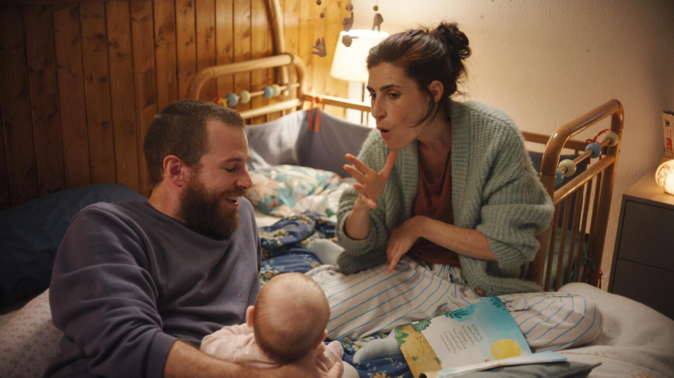 A man and a woman are sitting on a bed with a baby, engaging in an animated and warm interaction.