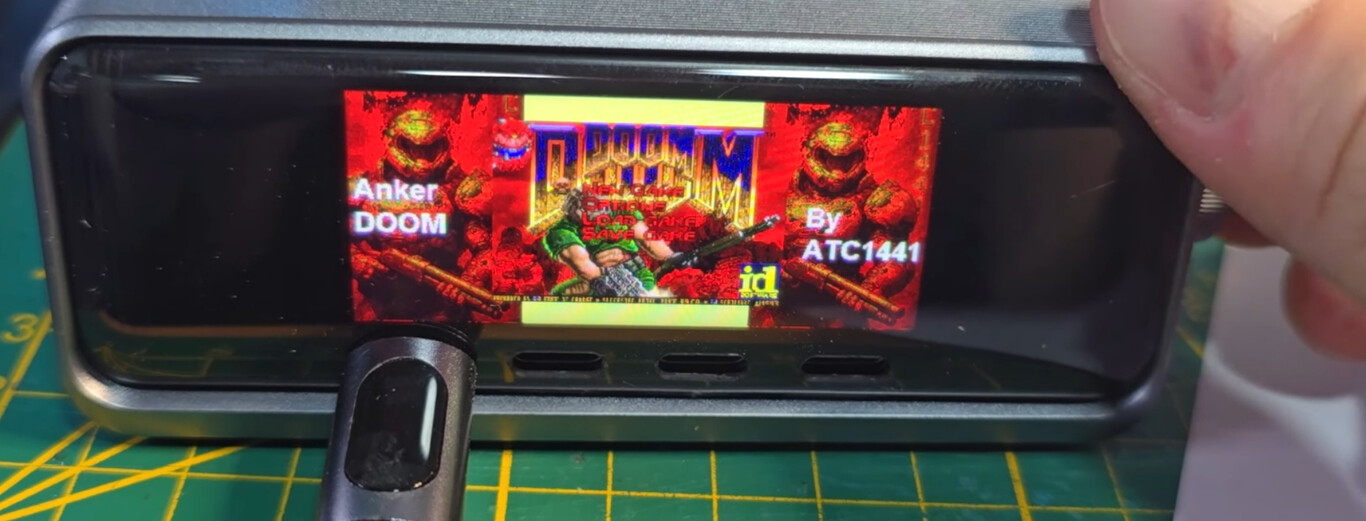 The image shows a close-up of a small rectangular digital screen encased in a black frame, resting on a green cutting mat with yellow grid lines. The screen displays the start menu of the classic video game "DOOM," featuring a central character in green armor holding a gun, set against a red and orange background with monsters and weapons. On the left side of the screen, the text "Anker DOOM" is visible, and on the right side, it says "By ATC1441." A person's fingers are slightly visible on the right edge of the frame, suggesting that the device is being held or adjusted.
