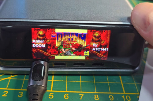 The image shows a small electronic device with a screen displaying the title screen of the video game "DOOM." The screen is surrounded by a black frame, and part of a person's hand is visible holding the device on the right side. A charging cable or power cord is plugged into the device on the left side, resting on a green cutting mat marked with yellow measurement lines and numbers. The game's title screen includes the word "Anker," "By ATC1441," and the iconic DOOM logo with artwork featuring a character and monsters.