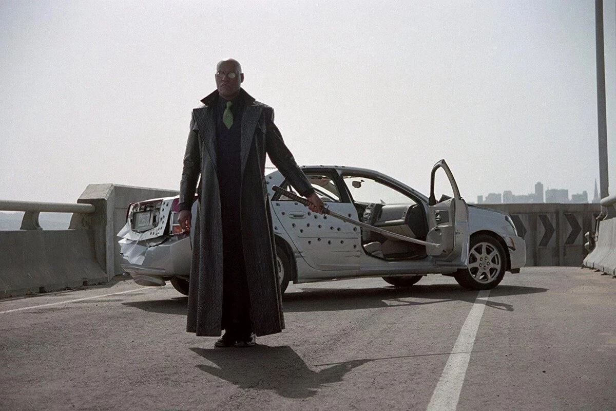 A man stands on a highway overpass next to a damaged silver sedan with its doors open. The car's side is riddled with bullet holes, and the rear bumper appears dented and bent out of shape. The man is wearing a long dark trench coat, a green tie, and reflective glasses, holding a katana sword loosely in one hand, casting a long shadow on the road beneath him. The background shows a hazy city skyline under a gray sky.