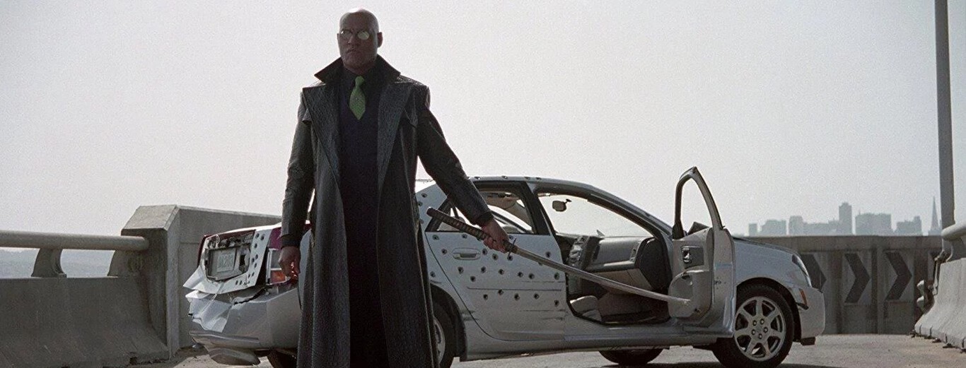 A bald man wearing dark sunglasses and a long black leather coat stands in front of a damaged silver car on a bridge. The car’s driver-side door is open, revealing the interior, and its side is riddled with numerous bullet holes. The man holds a large sword in his right hand, extending it downward, while a city skyline is visible in the hazy background.