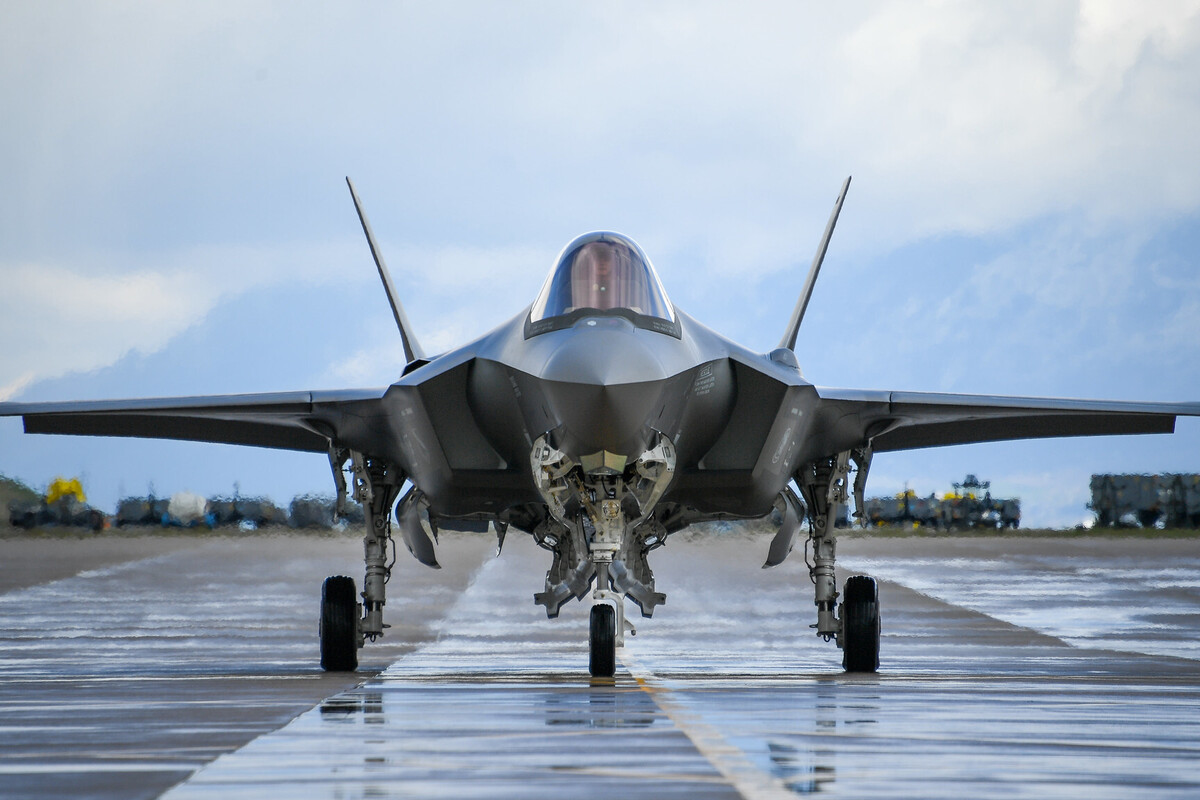 A modern stealth fighter jet is positioned on a wet runway, facing directly toward the viewer. The aircraft's sleek, angular design features twin vertical stabilizers and a single cockpit canopy with a reflective surface. Its landing gear is extended, and the background shows a cloudy sky with blurred shapes of vehicles or equipment along the horizon.