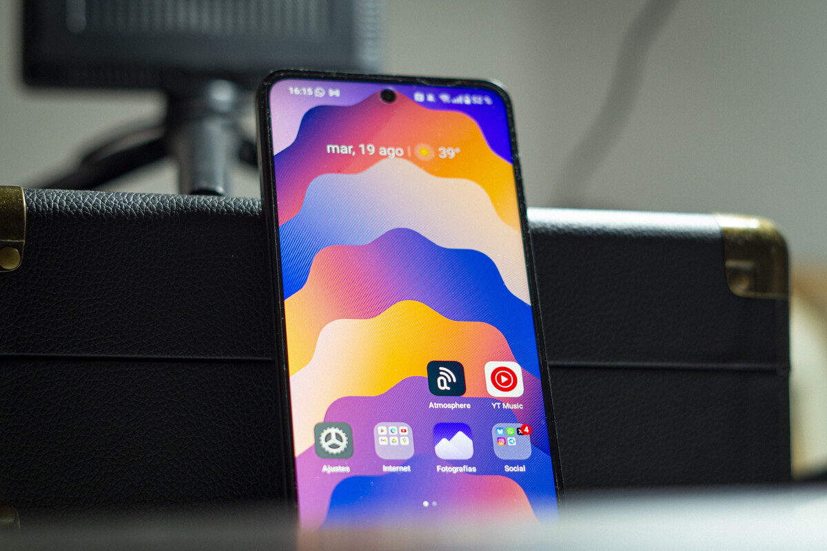 The image shows a smartphone standing upright against a black textured surface with brass corner protectors. The phone screen displays a colorful abstract wallpaper with wavy patterns in shades of purple, blue, orange, and yellow. The top of the screen shows the date 'mar, 19 ago' and a temperature of 39°C, along with the time '16:15.' Several app icons are visible near the bottom, including 'Atmosphere,' 'YT Music,' 'Ajustes,' 'Internet,' 'Fotografías,' and a 'Social' folder with notification badges. The background is softly blurred, focusing attention on the phone.