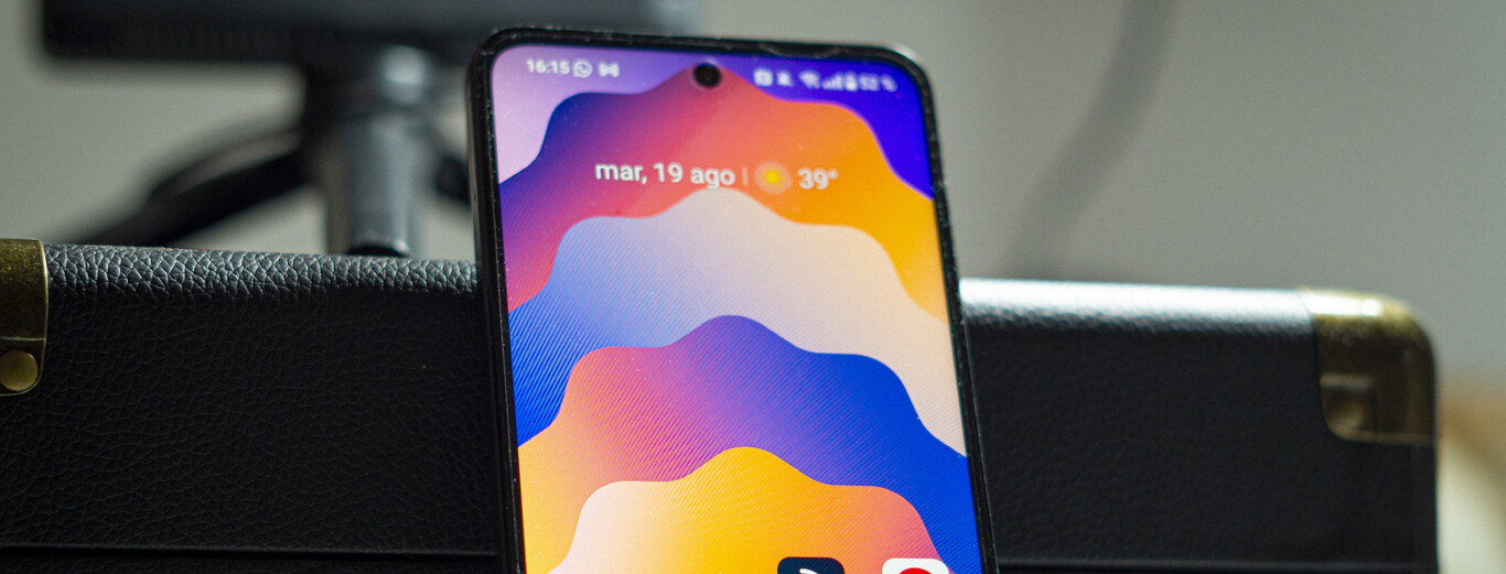 A close-up image shows the top portion of a smartphone screen resting against a black textured leather surface with brass corner protectors. The phone display features a colorful abstract wallpaper with wavy patterns in shades of orange, purple, blue, and yellow. At the top of the screen, the status bar indicates the time as 16:15, the date as Tuesday, August 19, and a weather icon showing a temperature of 39 degrees.