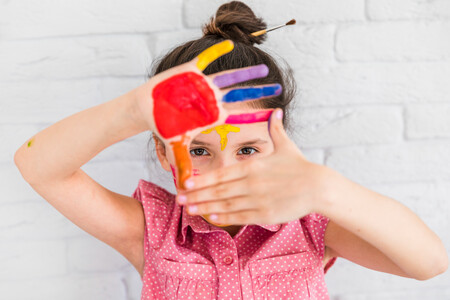 Portrait Of Girl Looking Through Her Painted Hands Standing Against White Brick Wall