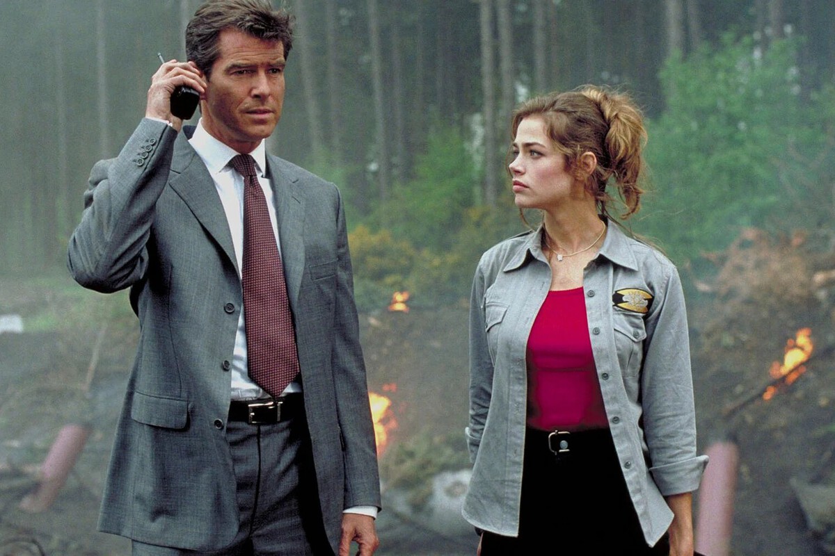 The image shows a man and a woman standing outdoors in a smoky, wooded area with small fires burning in the background. The man, dressed in a gray suit with a white shirt and red tie, holds a walkie-talkie or radio device up to his ear, appearing focused or serious. The woman, wearing a gray button-up shirt over a red top with a badge on her chest, looks intently at the man, her hair tied back in a loose ponytail. The scene suggests an intense or urgent situation, possibly related to law enforcement or emergency response.