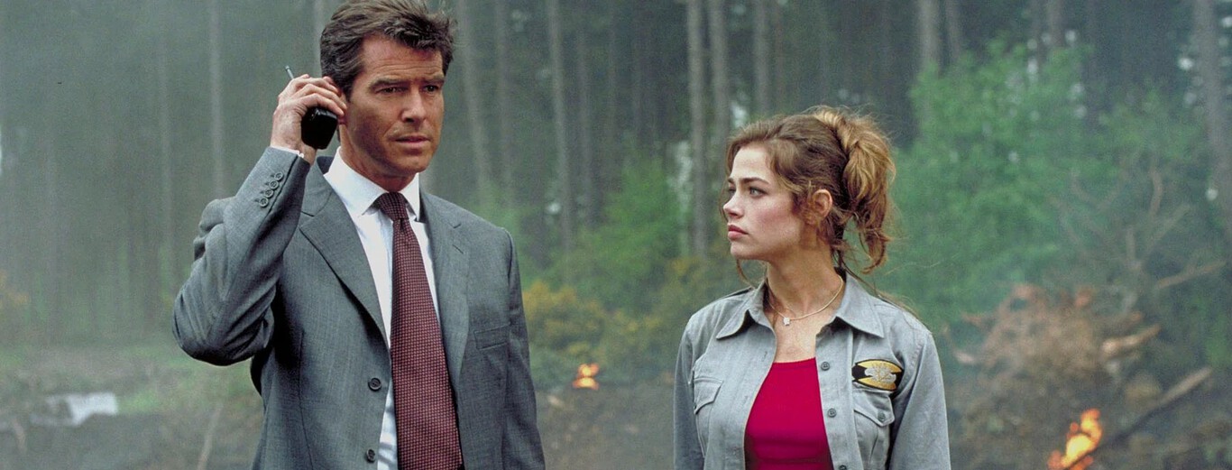 The image shows a man and a woman standing outdoors in a forested area with smoke and small flames visible in the background. The man, dressed in a gray suit with a white shirt and red tie, is holding a phone to his ear with a serious expression. The woman beside him wears a gray button-up shirt over a red top and looks thoughtfully to the side, her hair tied back loosely. The scene suggests a tense or urgent situation unfolding in a natural setting.