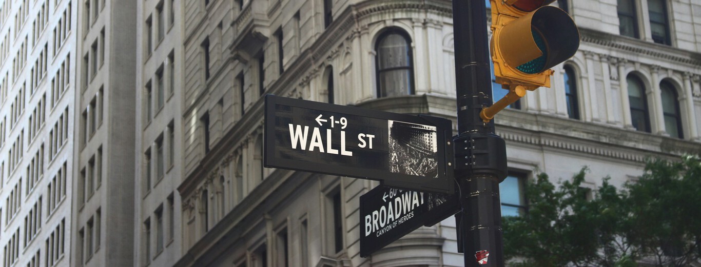 The image shows a street sign mounted on a black pole at an urban intersection. The sign prominently displays "WALL ST" with an arrow pointing left and numbers 1-9 above it, indicating the direction and address range. Below it, another sign indicates "BROADWAY" with a subtitle that reads "CANYON OF HEROES." In the background, there are tall, ornate stone buildings with many windows, characteristic of a historic financial district. A yellow traffic light is also visible, slightly to the right and above the signs, with trees partially visible in the lower right corner.