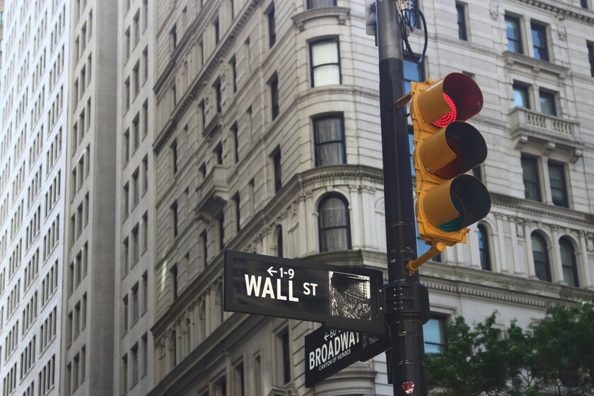 The image shows a street corner in a city with tall, historic stone buildings featuring large windows and classical architectural details. Prominently displayed is a black street sign for Wall Street, with an arrow pointing left and numbers 1-9 indicated, positioned above another sign for Broadway. A traffic light on the pole beside the signs is lit red, signaling vehicles to stop. The overall scene captures a busy urban environment with significant financial and cultural landmarks.
