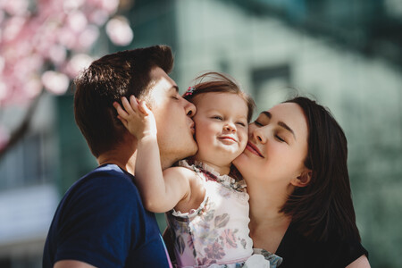 Happy Young Parents With Little Daughter Stand Under Blooming Pink Tree Outside