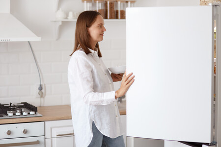 Retrato De Perfil De Hermosa Mujer Adulta Joven Con Camisa Blanca Mirando Sonriendo Dentro De La Nevera Con Una Sonrisa Agradable Sosteniendo El Plato En Las Manos Posando Con Cocina En Segundo Plano