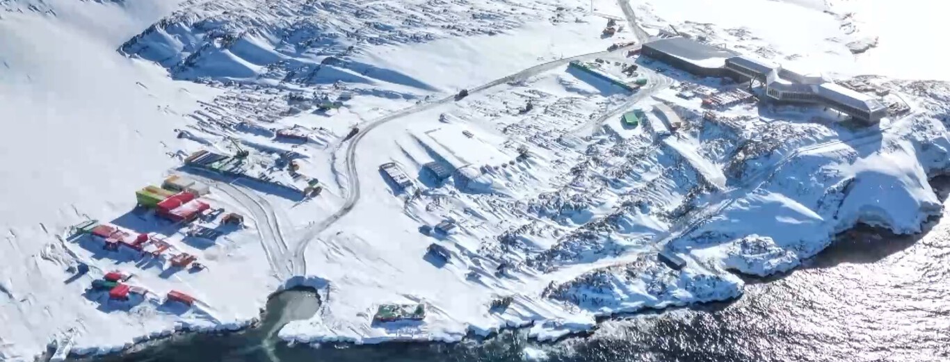 Snow-covered Antarctic coastal settlement with multiple buildings, colorful containers near water's edge.