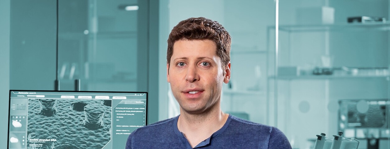 A man with short brown hair and blue eyes is standing in a modern laboratory setting. He is wearing a blue shirt and looking directly at the camera with a neutral expression. Behind him, there is a computer screen displaying a detailed scientific image labeled 'Double-stranded DNA' and some accompanying data, while the background shows glass walls and shelves with various lab equipment.