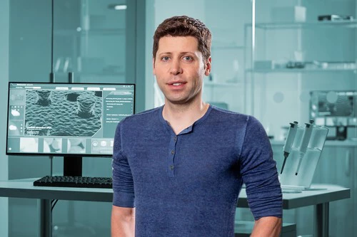 The image shows a man standing in a modern laboratory or research facility. He has short, curly brown hair and is wearing a blue long-sleeve shirt with a few buttons at the collar. Behind him is a computer monitor displaying highly magnified images resembling microscopic or nanoscale structures, indicating a scientific or technical environment focused on advanced research or analysis. The background includes glass walls and laboratory equipment, emphasizing a clean, high-tech workspace.