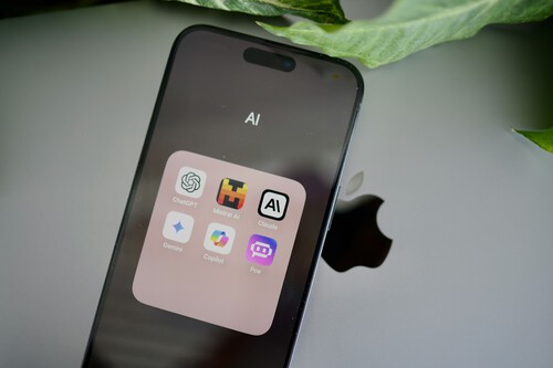 The image shows a smartphone displaying a folder labeled 'AI' on its screen. Inside the folder, there are six app icons arranged in two rows, including ChatGPT, Midjourney AI, Claude, Gemini, CoPilot, and Poe. The phone is resting on a silver laptop with a large, partially visible Apple logo, and a green leafy plant is in the background.