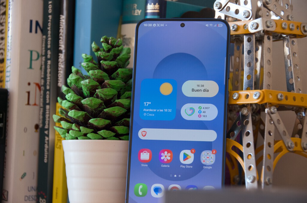 The image shows a smartphone with a blue-themed home screen displaying the weather, time, and several app icons, positioned upright on a surface. To the left of the phone, there is a white pot holding a large pinecone painted green on the scales. Behind the pinecone, a row of books is visible, with some titles in Spanish. On the right side of the phone, there is a metallic construction toy or model made of silver and yellow metal pieces with bolts. The overall setting appears to be a desk or shelf with a mix of natural elements, technology, and mechanical objects.