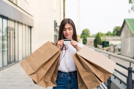 Mujer mirando con pena una tarjeta de crédito y cargada con muchas bolsas de compra de ropa