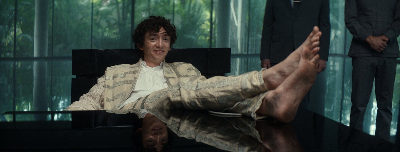 A young man with curly hair is sitting in a modern, black chair with his bare feet propped up on a reflective black table in front of him. He is smiling slightly and wearing a light-colored, patterned jacket over a white shirt and matching pants. Behind him, there are two people standing with their hands clasped in front of them, dressed in dark suits, and tall glass windows reveal a blurred green outdoor scene.