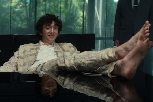 A young man with curly dark hair is sitting casually on a black, glossy surface, leaning back with a relaxed and confident expression. He is barefoot, with his legs stretched out in front of him and crossed at the ankles, wearing a cream and beige patterned suit over a white shirt. The background shows large glass windows with greenery visible outside, and a person dressed in a dark suit stands partially out of frame on the right side.