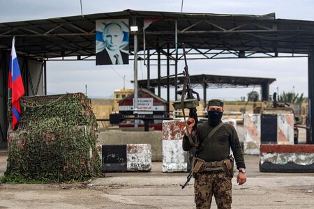 Un combatiente rebelde sirio se encuentra en un puesto de control de inspección para vehículos entrantes, frente a una bandera rusa y soldados rusos que custodian la entrada de la base militar siria de Hmeimim, arrendada por Rusia, en la provincia de Latakia, Siria, en 2024.