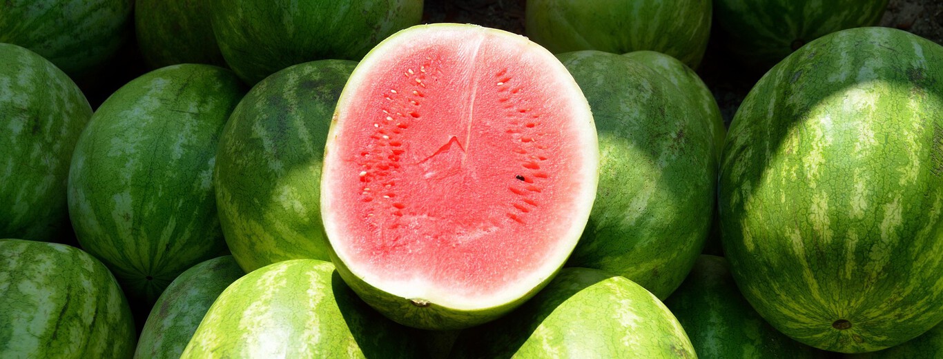 A group of whole watermelons with green, striped rinds is shown, with one watermelon cut in half and placed on top, revealing its bright red, juicy interior. The exposed flesh of the cut watermelon is dotted with small black and white seeds, highlighting its freshness. The watermelons are closely packed together, with sunlight casting soft shadows on their surfaces.