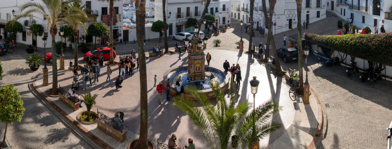 The image shows a sunlit town square with a circular fountain at its center, decorated with colorful tiles and some small plants. Surrounding the fountain are several tall palm trees casting long shadows on the ground. People are scattered around the square, some walking, others sitting on the benches along the edges, enjoying the pleasant atmosphere. White buildings with traditional architecture border the square, and cars are parked along the cobblestone streets. The overall scene conveys a lively yet relaxed urban space in a warm climate.