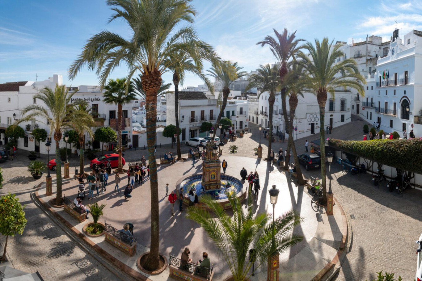 The image shows a sunny town square surrounded by white buildings with balconies and traditional architecture. Tall palm trees are scattered throughout the square, casting long shadows on the cobblestone ground. At the center of the square is a decorative fountain, around which people are gathered, some walking, sitting on benches, or socializing. The atmosphere appears lively and relaxed, with a clear blue sky overhead.